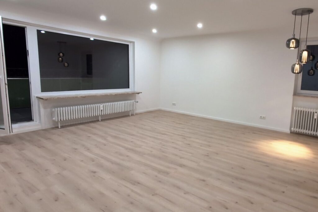 Bad Harzburg: living room after the renovation, in the evening: large window and balcony door, second window with a pendant light, radiators and light wood-look flooring, February 2026
